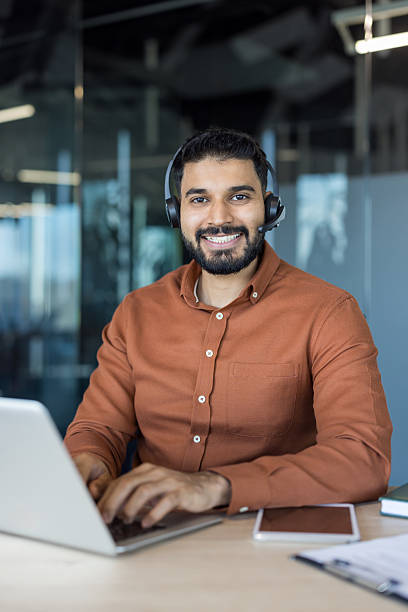 Smiling male call center agent wearing a headset, providing online assistance and technical support while typing on a laptop in a modern corporate office environment